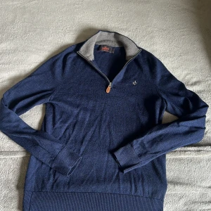 Marinblå merinoulltröja från Morris - Säljer en marinblå långärmad tröja från Morris i 100% merinoull. Tröjan har half zip med läderdetalj, ribbade muddar och en broderad logga på bröstet. Perfekt för dig som gillar klassisk och stilren look.