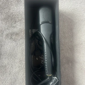 GHD svart plattång - Snygg svart plattång från GHD med guldiga detaljer. Ergonomiskt handtag med prickig textur för bättre grepp. Perfekt för att styla håret rakt eller skapa lockar. Kommer med sladd och originalförpackning.