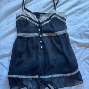 Svart linnetopp med spets och knappar - Supersöt svart linnetopp med tunna axelband, vit/beiga spetsdetaljer och knappar framtill. Toppen är luftig och har en lätt volang nertill.