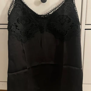 Svart spetslinne från H&M XS - Supersnygg svart linnetopp från H&M i storlek XS. Toppen har tunna, justerbara axelband och vacker spetsdetalj framtill med blommigt mönster. Materialet är glansigt och känns lyxigt.