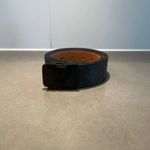 Svart Louis Vuitton skärp i skinn - Snyggt svart skärp från Louis Vuitton med klassiskt LV-spänne i svart metall. Bältet har diskret monogrammönster och är gjort i äkta skinn med brun insida. Perfekt accessoar för att lyfta din outfit med en lyxig touch.