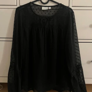 Svart prickig blus från Vila, stl 34 - Säljer en svart blus från Vila i storlek 34. Blusen har transparenta, långärmade ärmar med prickigt mönster och en söt knytning vid halsen. Den är i nyskick och aldrig använd.