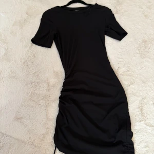 Svart kort klänning från H&M Divided - En svart, tight kortklänning från H&M Divided i storlek XS. Klänningen har korta ärmar och snygg rynkdetalj på sidorna med snörning. Enkel och stilren modell som sitter figurnära och är perfekt för dig som gillar minimalistisk stil.