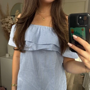 Blåvit offshoulder klänning med volang - Säljer en söt blåvit randig klänning med offshoulder och volangdetalj upptill. Perfekt för varma dagar och har en luftig passform. Materialet känns lätt och somrigt.