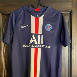 PSG matchtröja  - Paris Saint-Germain fotbollströja i mörkblått med röd och vit rand längs mitten. Klassisk PSG-logga på bröstet, Nike Swoosh och sponsortryck. Tillverkad i Dri-FIT-material som andas. Kortärmad modell, perfekt för fotboll eller supporterliv.