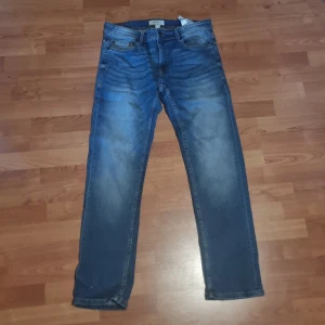 Blå jeansbyxor 33/32 - Säljer ett par klassiska blå jeans från Jack & Jones i storlek 33/32. Jeansen har en rak passform med snygga slitningar och ljusare partier framtill. Tillverkade i bomull med fem fickor och orangea sömmar. Perfekta för en avslappnad stil.
