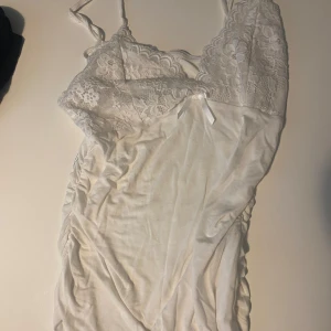 Vit spetsklänning med rosett - Supersöt vit topp  med tunna axelband, spets upptill och liten rosett framtill. Toppen  har omlottkänsla och är figurnära med rynkade sidor. 