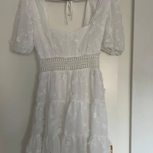 Vit blommig kortklänning med spets - Superfin vit kortklänning med broderade blommor, puffärmar och markerad midja med spetsdetaljer. Klänningen har omlottknytning i ryggen och volang i kjolen. 