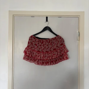 Röd volangkjol med mönster - Superfin kort röd kjol med vitt grafiskt mönster och dubbla volanger. Kjolen har elastisk midja och är gjord i ett lätt, luftigt material som känns perfekt för varmare dagar. Snygg att styla med en enkel topp och sneakers.