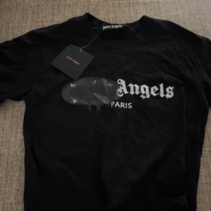 Svart Palm Angels t-shirt med tryck - Svart t-shirt från Palm Angels med ikoniskt Paris-tryck framtill och coolt spray paint-detalj över loggan. Klassisk passform och rund hals. Perfekt för dig som gillar streetwear och vill sticka ut med en snygg statement tee.