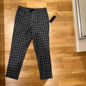 Gråsvarta rutiga byxor New Look S - Snygga gråsvarta byxor från New Look i storlek S. Byxorna har rutigt mönster, dragsko i midjan och croppad längd. Perfekta för dig som gillar en stilren look med lite edge. Materialet känns mjukt och bekvämt, passar till många outfits.