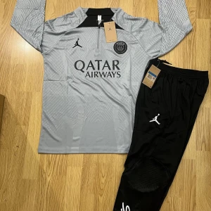 PSG långärmad träningströja Jordan - Snygg långärmad PSG träningströja i ljusgrått med svarta detaljer och subtilt geometriskt mönster. Jordan-logga och PSG-emblem på bröstet, samt Qatar Airways-tryck. Materialet är lätt och andas, perfekt för fotbollsträning.