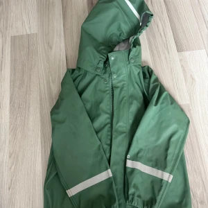 Grön regnjacka från Reima 116 cm - Grön regnjacka från Reima med huva och reflexdetaljer på ärmar och huva. Jackan har dragkedja framtill, tryckknappar och elastiska muddar. Perfekt för blöta dagar och håller dig torr oavsett väder. Snygg och funktionell design för aktiva dagar ute.