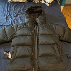Svart pufferjacka från Everest - Säljer en svart pufferjacka från Everest med huva och dragkedja. Jackan är riktigt fluffig och har en clean look med breda kanaler. Perfekt för kalla dagar när du vill hålla värmen och ändå se stilren ut. Passar dig som gillar enkel och sportig stil.