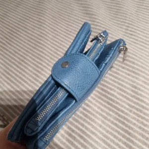 Blå plånbok från Misako i skinn - Snygg blå plånbok från Misako med flera kortfack, fönster för ID och två dragkedjefack. Stängs med tryckknapp och har stilren, modern design. Perfekt för dig som vill ha ordning på allt i väskan.