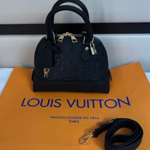 Svart Louis Vuitton liknande handväska i skinn - Säljer en svart handväska med klassiskt monogrammönster i äkta skinn. Väskan har guldfärgade metalldetaljer, dubbla handtag, avtagbar axelrem och dragkedja. Samt nyckel och lås så man kan låsa väskan