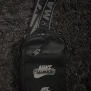 Nike Air Max svart axelväska - Svart axelväska från Nike Air Max med justerbart band där det står 'Air Max' i vitt. Väskan har dragkedja upptill och framtill, samt tydlig Nike-logga på framsidan. Perfekt för att bära det viktigaste och passar till streetwear.