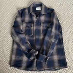 Rutig overshirt från Dobber i blått - Snygg rutig overshirt från Dobber i mörkblå och beige toner. Skjortan har två bröstfickor med lock och knappar framtill. Perfekt lager-på-lager-plagg med lång ärm och klassisk krage. Mjuk och lite tjockare känsla, passar till många stilar.