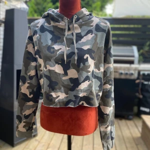 Camouflage croppad hoodie från H&M Divided M - Trendig croppad hoodie med ett coolt camouflagemönster. Den har en avslappnad passform och en rå kant nedtill, vilket ger den en modern och edgy look. Perfekt för en avslappnad men stilfull outfit. Längd: 45 cm ﻿Bystvidd: 60 cm ﻿60% bomull, 40% polyester, 5% elastan  Inga bilder när den sitter på mig! Katter finns i hemmet!