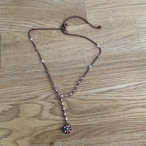 Rosa halsband med blomma och pärlor - Sött halsband i roséguld med små rosa stenar och vita pärlor längs kedjan. Hängsmycket är format som en blomma med rosa och turkosa detaljer. Justerbar längd med knyt baktill. Perfekt för dig som gillar romantiska accessoarer.
