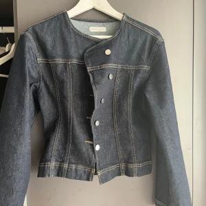 Mörkblå jeansjacka från Gina Tricot  - Snygg mörkblå jeansjacka med asymmetrisk knäppning och vita knappar. Jackan har raka linjer, rund halsringning och kontrastsömmar. Aldrig använd storlek S/34 