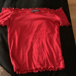 Röd offshoulder topp Brandy Melville - Superfin röd offshoulder topp från Brandy Melville med ribbad struktur och vågiga kanter. Toppens tighta passform och korta ärmar ger en snygg och trendig look. Perfekt att styla med jeans eller kjol för en cool vibe.