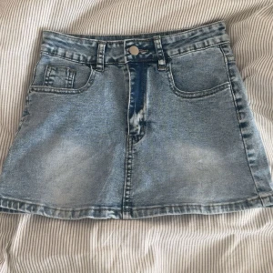 Ljusblå jeanskjol i kort modell XS - Snygg ljusblå jeanskjol i kort modell med klassiska fickor fram och bak samt hällor för bälte. Kjolen har en lätt tvättad look och stängs med knapp och dragkedja. Perfekt för dig som gillar en avslappnad och trendig stil. Den är aldrig använd 