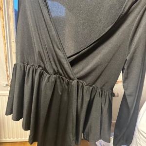 Svart omlottopp från SHEIN Curve - Svart omlottopp från SHEIN Curve i storlek 1XL. Toppen har långa ärmar, v-ringning och volangdetalj nedtill som ger en snygg och flowy look. Tillverkad i ett mjukt och lätt material som känns skönt mot huden.