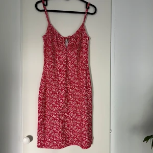 Röd blommig kortklänning med knyt - En söt röd klänning med vita blommor. Passar perfekt till sommaren. Klänningen har en även en smickrande detalj vid bysten med en snörning. 