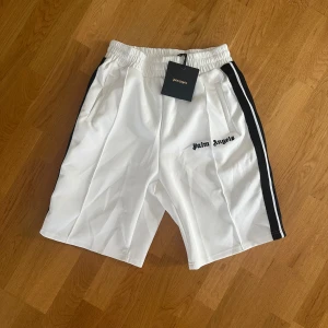 Vita Palm Angels shorts med svarta ränder - Snygga vita shorts från Palm Angels med svarta sidoränder och broderad logga på vänster ben. De har elastisk midja och sidofickor, perfekta för en sportig och trendig look. Materialet är mjukt och känns bekvämt mot huden. Storlek M