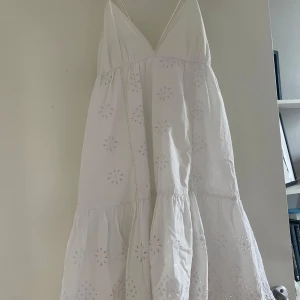 Vit somrig klänning med broderi - Superfin vit klänning med tunna axelband och v-ringning. Klänningen har broderade blommor och volangkant nertill. Den är ärmlös och har knytning i ryggen. Perfekt för varma dagar och ger en riktigt fräsch vibe.  Lite missfärgad.
