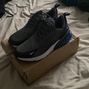 Nike Air Max 270 - Nike Air Max 270 sneakers i svart och mörkgrå mesh med blå detaljer och stor synlig Air Max-enhet i hälen. Skorna har snörning, rund tå och vit sula. Perfekta för dig som gillar sportig och modern stil.