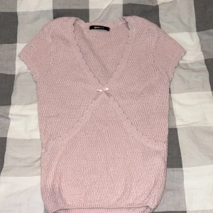 Ljusrosa stickad omlottopp Gina Tricot - Supersöt ljusrosa stickad omlottopp från Gina Tricot med korta ärmar och volangkant längs v-ringningen. Toppen har en liten rosett framtill och är croppad i modellen. Perfekt för dig som gillar gulliga detaljer och mjuka pasteller.
