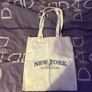 Vit tygkasse med New York-tryck - Vit shoppingväska i bomull med tryck i blått och svart där det står 'NEW YORK MANHATTAN'. Väskan har långa handtag och är perfekt att slänga över axeln när du ska ut på stan. Enkel och stilren design som passar till det mesta.