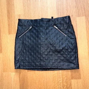 Svart quiltad skinnkjol med dragkedjor - Snygg svart kortkjol i quiltat skinnimitation med två silvriga dragkedjor framtill. Kjolen har en cool rutstruktur och sitter tajt för en edgy look. Perfekt att styla med boots eller en oversized jacka.