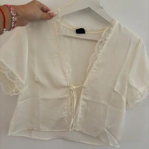 Vit omlottblus med spets från Gina Tricot - Supersöt vit omlottblus från Gina Tricot i glansigt material med spetsdetaljer längs kanterna och ärmsluten. Knytband framtill och kort ärm. Superfin😍😍