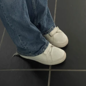 Vita sneakers från J.Lindeberg - Snygga vita sneakers från J.Lindeberg med kardborreband och platt sula. Skorna har en clean look med rund tå och är tillverkade i syntetmaterial. Perfekta för dig som gillar stilrena skor.