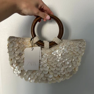 Slutsåld väska Zara - Superfin beige handväska från Zara med stora trähandtag och massor av skimrande pärlpaljetter på utsidan. Väskan har ett innerfack med dragkedja och magnetknäppning. 