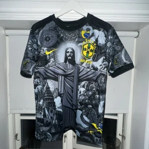 Brasil Nike fotbollströja svart/gul - Unik Brasilien fotbollströja med svart och grått konstnärligt tryck framtill, stora gula detaljer och CBF-emblem. Kortärmad modell i lätt och ventilerande material, perfekt för dig som vill sticka ut på planen eller läktaren.