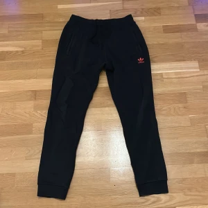 Svarta Adidas mjukisbyxor med röd logo - Svarta mjukisbyxor från Adidas med röd broderad trefoil-logo och text på vänster lår. Byxorna har resår i midjan, snörning och muddar vid bensluten. Perfekta för chill eller träning, med två fickor framtill.