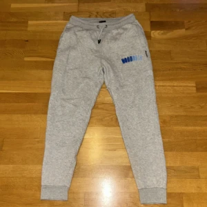 Grå mjukisbyxor från Hoodrich - Snygga grå mjukisbyxor från Hoodrich med blå logga på ena benet. Byxorna har snörning i midjan, muddar vid bensluten och är tillverkade i mjukt bomullsmaterial. Perfekta för chill eller träning.