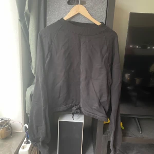 Svart croppad tröja från Weekday - Svart croppad sweatshirt från Weekday med långa ärmar och snörning i nederkanten. Tröjan har en relaxed fit och är tillverkad i mjuk bomull. Perfekt för en chill och stilren look.