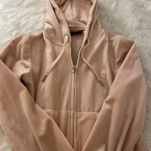 Beige rosa velourjacka Juicy Couture - Supermjuk och snygg beige rosa velourjacka från Juicy Couture med huva, dragkedja och snörning. Klassisk JC-logo broderad på bröstet. Perfekt för en chill och trendig look.