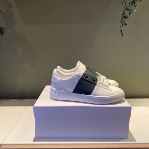 Valentino Garavani Open sneakers vit/svart - Snygga Valentino Garavani Open sneakers i vitt skinn med bred svart rem över vristen. Klassisk rund tå, vita snören och ikoniska nitar på hälen. Kommer med originalpåse och box. Perfekta för dig som gillar stilrena och lyxiga detaljer.  Følger med alle tilbehør!!