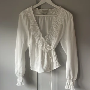 Vit omlottblus från Neo Noir - Supersnygg vit omlottblus från Neo Noir med långa ärmar och volangkant längs v-ringningen. Blusen har knyt i sidan och smockade ärmslut för en extra touch. Materialet är glansigt och känns lyxigt, perfekt för att lyfta vilken outfit som helst.💕den är i s/m 