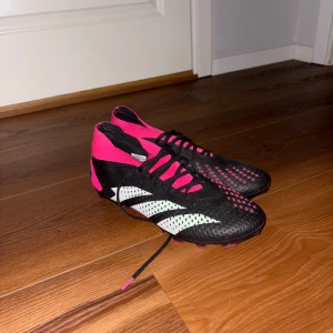 Adidas Predator Svart/Rosa Fotbollsskor - Snygga Adidas Predator fotbollsskor i svart med rosa och vita detaljer. Skorna har snörning, högre strumpliknande skaft och mönstrad ovandel för extra grepp på planen. Perfekta för dig som vill sticka ut med stil och prestanda på matchen. Sitter även lite litet men passar perfekt 44