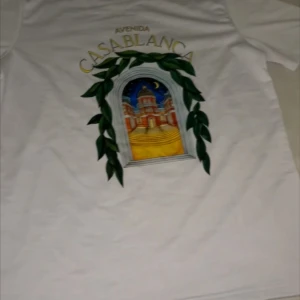 Vit t-shirt med Casablanca-tryck - Vit t-shirt med färgglatt tryck av en byggnad och texten 'Avenida Casablanca' på ryggen. Trycket har gröna blad och en nattlig himmel. Klassisk passform och korta ärmar, perfekt för en chill look.