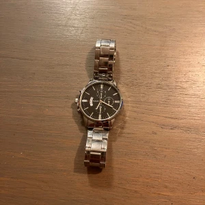 Svart och silver klocka  - Stilren klocka  med svart urtavla och silverfärgat armband i metall. Klockan har flera små urtavlor, dag- och datumvisning samt klassisk rund form. Perfekt accessoar för dig som gillar en clean och tidlös look.