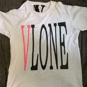 Vit VLONE t-shirt med rosa tryck - Snygg vit t-shirt från VLONE med stort svart och rosa logotryck på framsidan och en stor rosa V på ryggen. Klassisk passform och rund hals. Perfekt statement-plagg för streetwear-stilen.