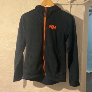 Svart fleecejacka från Helly Hansen - Svart fleecejacka från Helly Hansen med orange dragkedja och HH-logga på bröstet. Jackan har huva och långa ärmar, perfekt för kyliga dagar. Mjuk och skön känsla, passar dig som gillar en sportig och avslappnad stil.